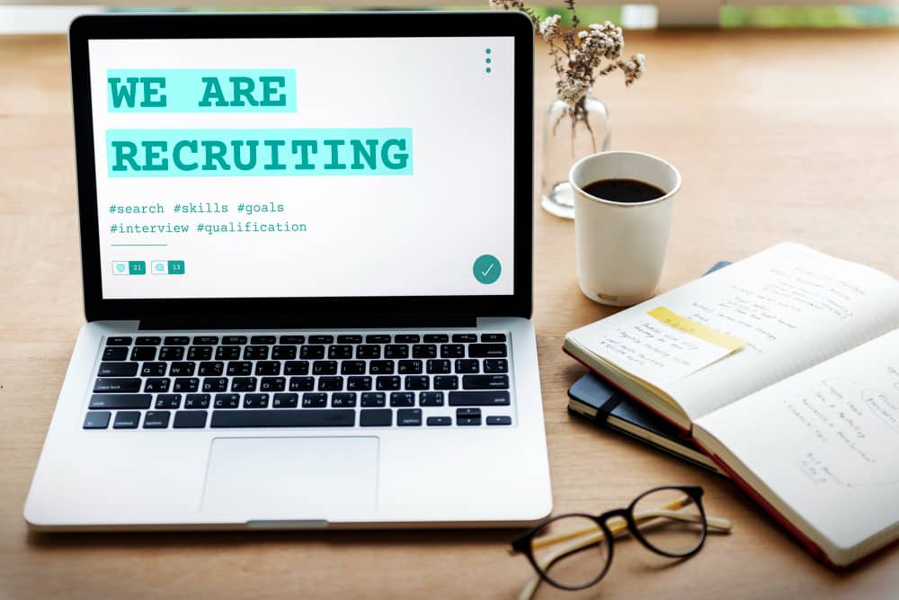 Laptop displaying "We Are Recruiting" with relevant hashtags, placed beside a notebook, coffee cup, and eyeglasses, indicating a workspace for recruitment activities.