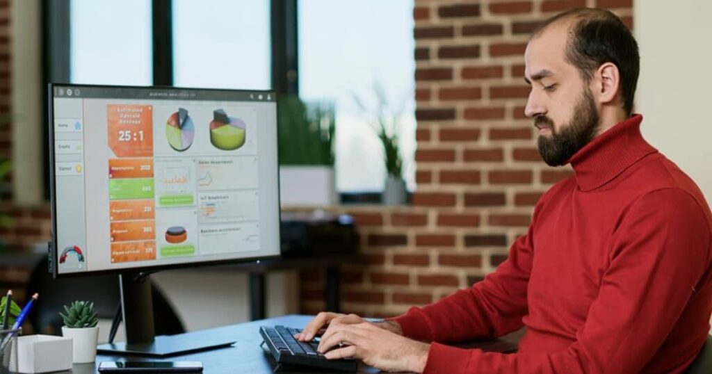 A man in a red turtleneck working on a desktop computer displaying business analytics and data visualization charts.