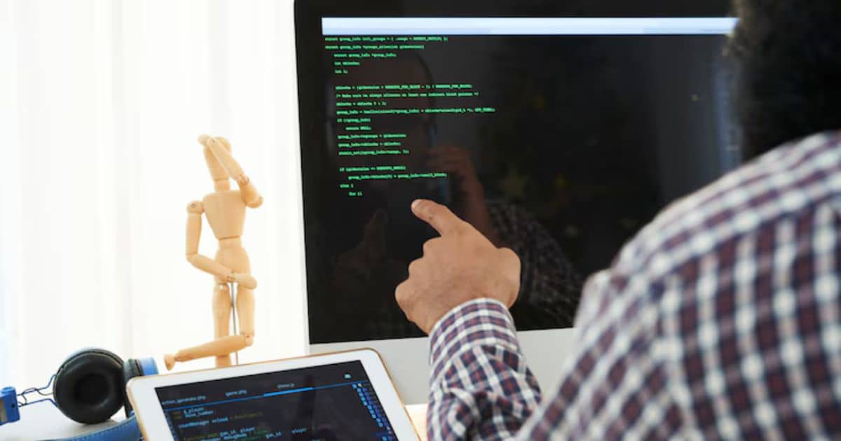 A developer pointing at code displayed on a large monitor with a tablet showing additional code on the desk, accompanied by a wooden mannequin figurine.