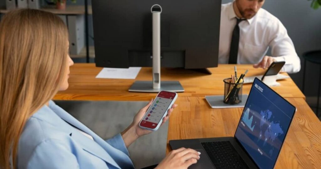 A businesswoman using a mobile phone and laptop, with a colleague working on a phone in the background.