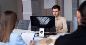 A focused man working at a computer during a recruitment interview, with two interviewers visible in the foreground.